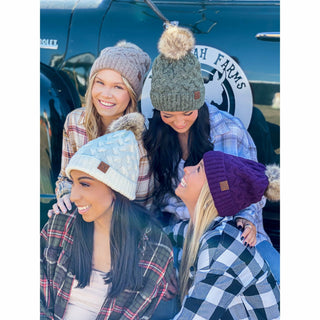 Four women wearing cable knit CC beanies with faux fur poms in taupe, green, ivory, and purple