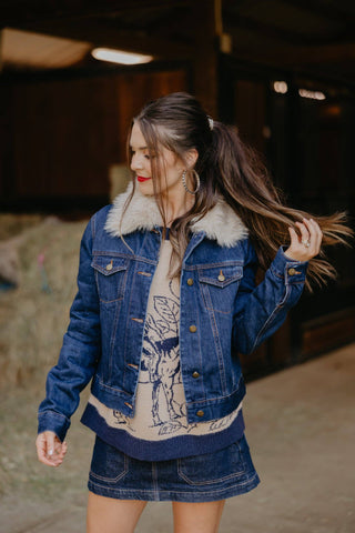 Woman wearing a denim jacket with fur trim and a graphic t-shirt, standing outdoors.