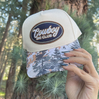 Tan snapback hat with Cowboy Ski Club embroidered patch and snowy treeline camo brim