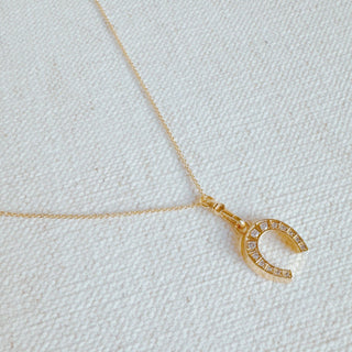 A gold-colored pendant necklace with a horseshoe shape adorned with small sparkling stones, displayed against a white fabric background.