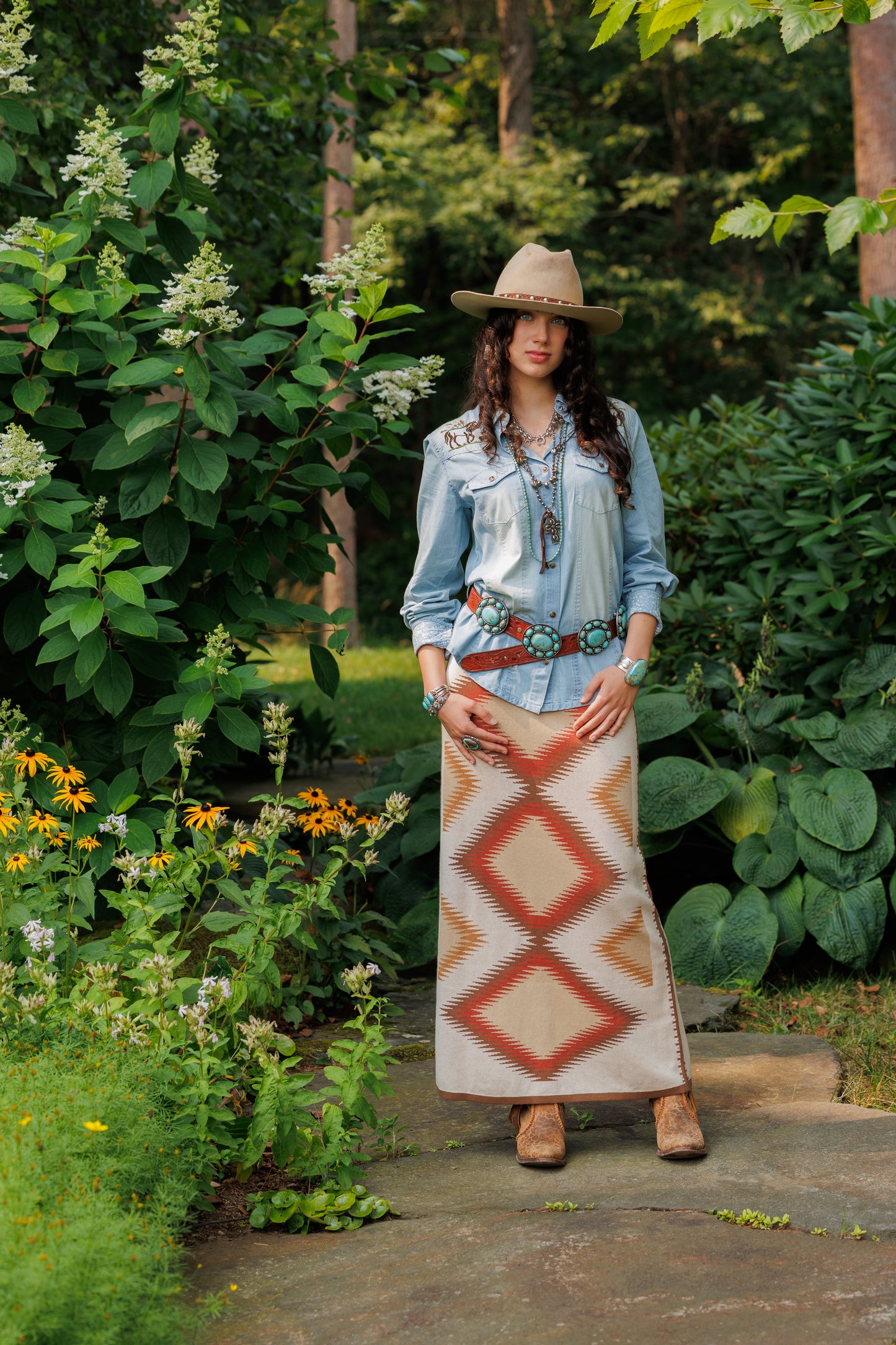 Woman in a denim jacket and patterned skirt standing in a garden with greenery and flowers.