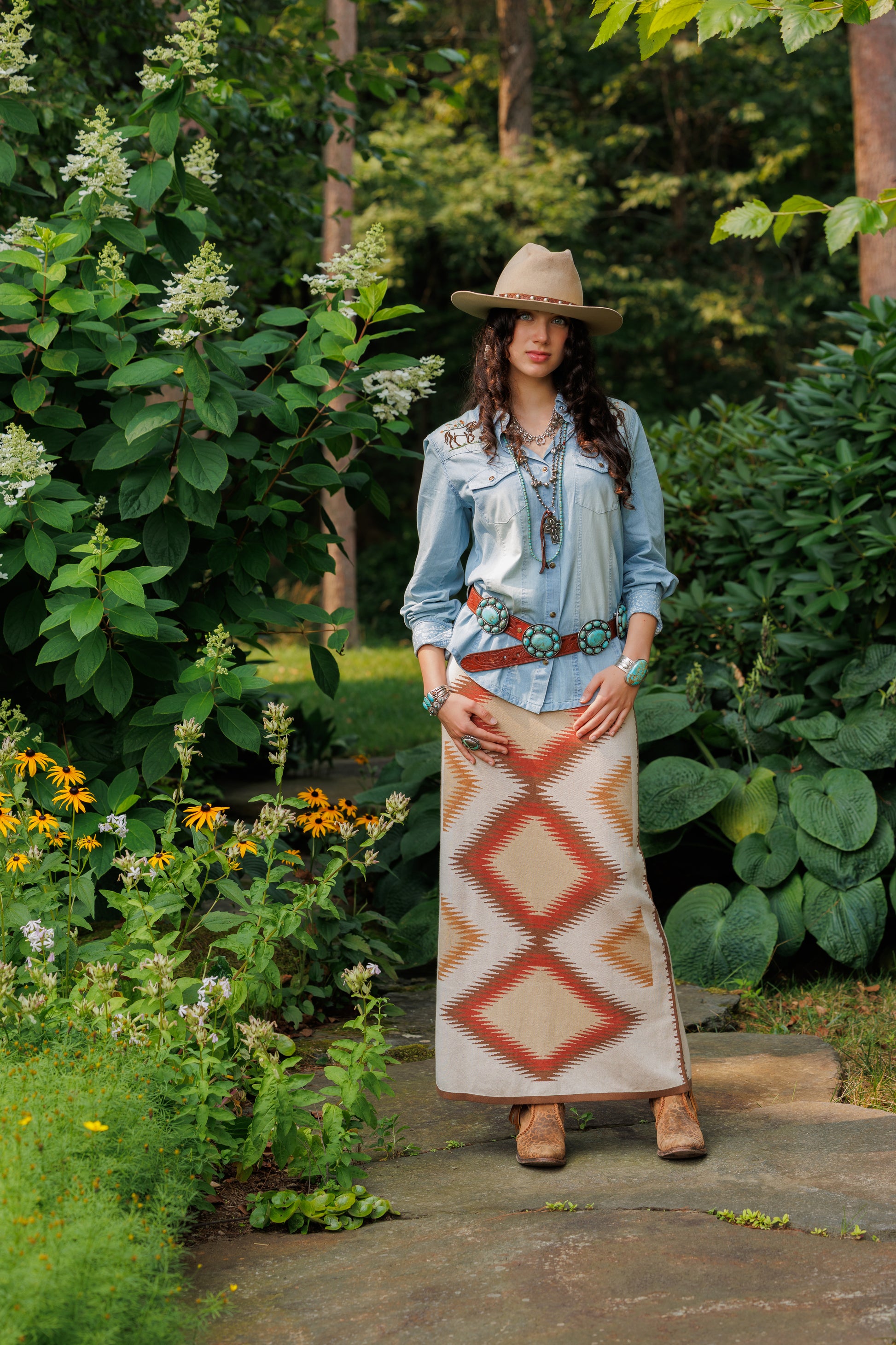 Woman in a denim jacket and patterned skirt standing in a garden with greenery and flowers.