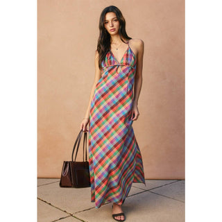 Woman wearing a colorful plaid dress holding a brown handbag against a beige wall.