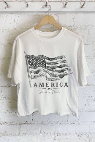 White t-shirt with American flag graphic and 'America 250 Years of Freedom' text on a hanger against a white brick wall.