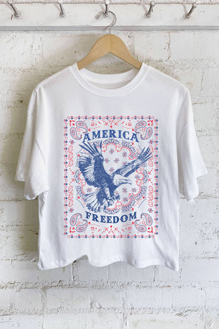 White t-shirt with 'America Freedom' graphic on a wooden hanger against a white brick wall.