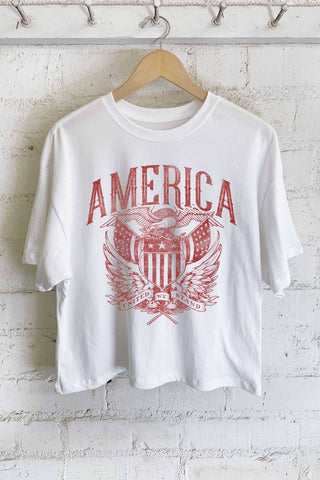 White t-shirt with red 'AMERICA' graphic on a hanger against a white brick wall.