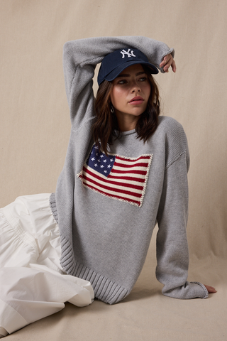 Woman wearing a gray sweater with an American flag patch and a blue cap, sitting on a beige background.