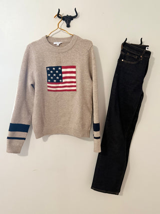 Beige sweater with American flag design and black jeans on a beige wall.