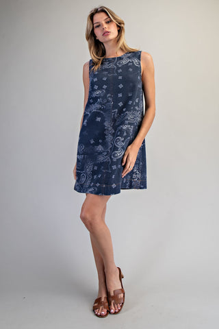 A woman modeling a sleeveless navy blue shift dress with a white paisley and floral bandana print, paired with brown leather slide sandals, against a neutral gray studio background.