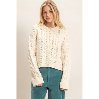 A woman wearing a cream cable-knit cropped sweater with a buttoned neckline and teal corduroy pants, standing against a plain light beige studio background.