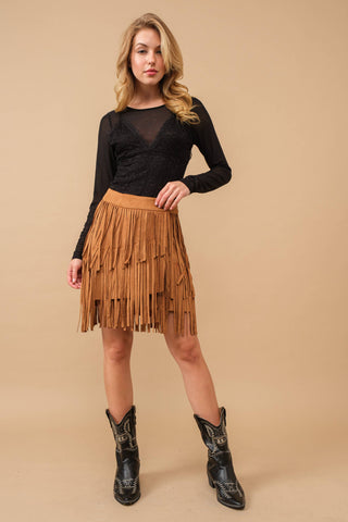 Woman wearing a black top and brown fringe skirt with black boots on a beige background