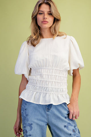 A woman wearing a white short-sleeve blouse with puff sleeves and a gathered smocked waist, paired with light blue jeans, against a solid light green background.