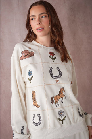 A woman wearing a cream-colored crewneck sweatshirt with western-themed embroidered patches including a cowboy hat, boots, horseshoes, a horse, and flowers, set against a textured mauve background.