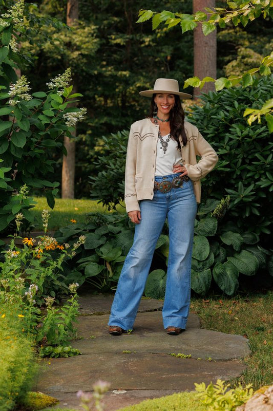 Woman in a beige jacket and blue jeans standing on a garden path with greenery around.