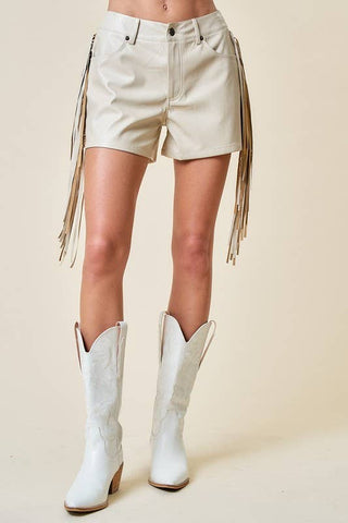 A model's lower body wearing off-white leather fringe shorts and matching off-white leather cowboy boots, set against a solid beige studio background.