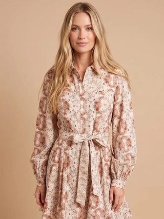 A woman modeling a long-sleeve button-front dress with a tie waist and an allover paisley print in warm beige, brown, and muted red tones, against a solid beige studio background.