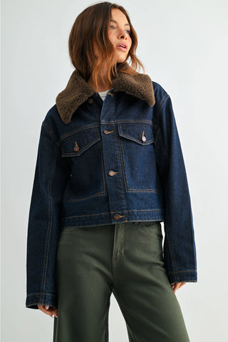 A woman wearing a cropped dark blue denim jacket with a brown shearling-style collar, paired with olive green pants, photographed against a light gray studio background.