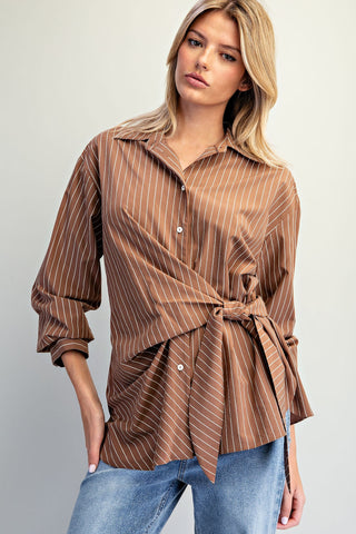 A woman wearing a brown and beige vertically striped wrap-style button-up shirt with a tie waist, paired with blue jeans, against a plain light gray background.