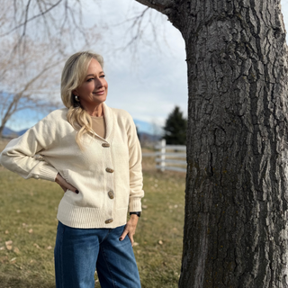 Woman in a cream cardigan and blue jeans standing next to a tree in an outdoor setting.