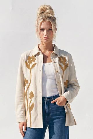 A woman modeling a cream-colored button-up shirt jacket with tan floral embroidery, worn over a white ribbed tank top and blue jeans against a plain white background.
