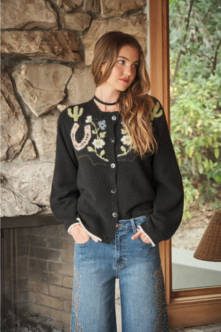 A woman wearing a black knit cardigan with embroidered floral and western motifs, paired with blue jeans, standing in front of a stone fireplace and window.