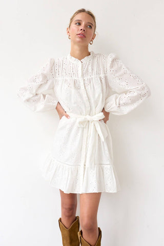 Woman wearing a white dress with long sleeves and a belt, standing against a white background.