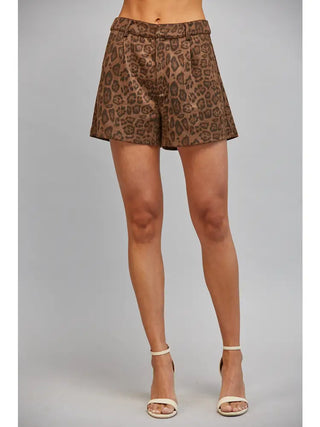 A model's lower body wearing brown and beige leopard-print shorts and cream high-heeled sandals, standing against a solid gray studio background.