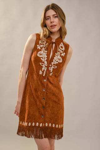 Woman wearing a brown suede dress with white embroidery and fringes on a beige background