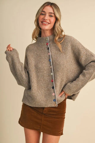A woman modeling a cozy, oversized taupe knit sweater with colorful decorative buttons down the front, paired with a brown mini skirt, against a solid beige background.