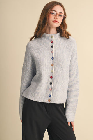 A woman wearing a light gray textured knit mock-neck sweater with a vertical row of colorful buttons down the front, paired with black pants, against a solid beige background.