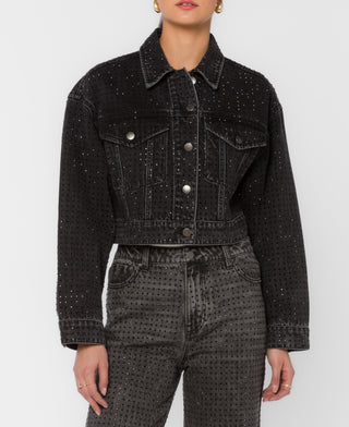 A model wearing a cropped black denim jacket with silver rhinestone embellishments, paired with matching black denim jeans, against a light gray studio background.