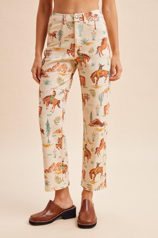 A model is shown from the waist down wearing high-waisted, cropped pants with a western-themed print of horses, cacti, and riders on a cream background, paired with brown leather mule shoes against a solid beige backdrop.