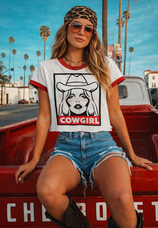 A woman wearing a white ringer t-shirt with red trim and a black-and-white cowgirl graphic that reads 'COWGIRL' in red, sitting in the bed of a red pickup truck outdoors.