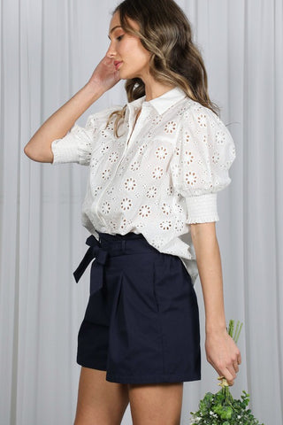 Woman wearing a white blouse with floral patterns and navy shorts against a light background