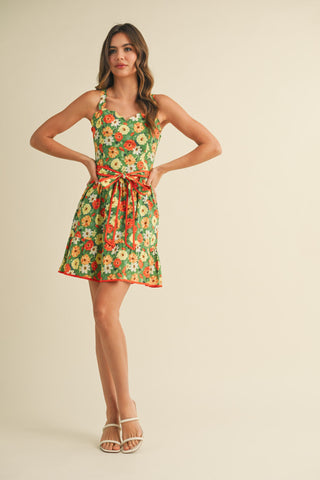 A model wearing a short green floral dress with scalloped edges and a contrasting belt, standing against a neutral background.