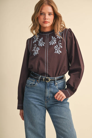 Model wearing a dark brown long-sleeve mock neck blouse with light blue floral embroidery and ruffled neckline