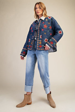 A woman models a dark blue quilted jacket with colorful floral embroidery, paired with light-wash cuffed jeans and tan ankle boots, against a solid beige studio background.