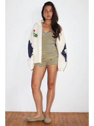 A woman stands against a white wall wearing a yellow and blue striped romper, a cream knit cardigan with blue lace sleeves and a green rose patch, and taupe slip-on shoes.
