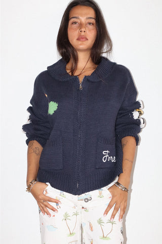 A woman modeling a navy blue knit zip-up hoodie with a green embroidered patch, white embroidered script on the pocket, and white patterned pants against a plain white background.