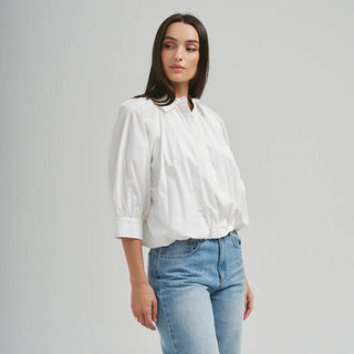 A woman wearing a white long-sleeve blouse with gathered puff sleeves and light blue jeans, standing against a plain light gray studio background.