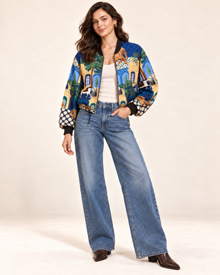 A woman modeling a vibrant, multicolored patterned bomber jacket with blue wide-leg jeans and brown ankle boots, standing against a plain beige studio background.