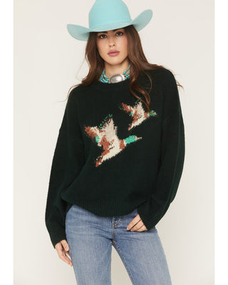 Woman wearing a black sweater with star design, light blue jeans, and a turquoise cowboy hat on a white background.