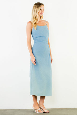 A woman modeling a light blue sleeveless midi dress with thin straps and a square neckline, standing against a plain white wall.
