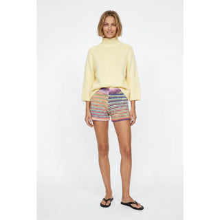 A woman modeling a pale yellow mock-neck knit sweater and multicolored striped knit shorts against a plain light gray studio background.