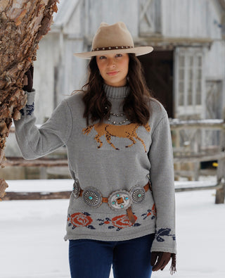 A woman wearing a gray knit sweater with a brown bison and southwestern-style geometric patterns, styled with a tan hat, belt, and blue jeans, standing outdoors next to a tree in a snowy setting.