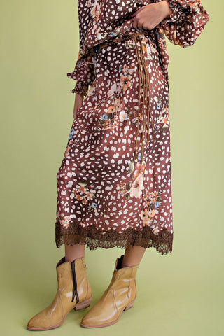 A person wearing a brown floral midi dress with white polka dots and tan ankle boots, standing against a solid light green background.