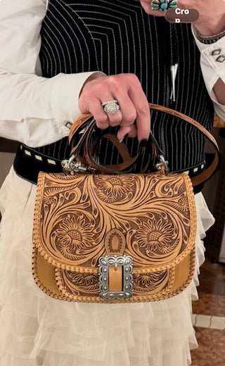 A close-up of a hand holding a tooled leather shoulder bag. The bag is tan and brown with intricate floral tooling and a silver-toned concho-style buckle.
