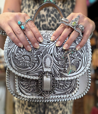 Hands holding a floral-tooled leather handbag with silver hardware and whipstitch trim