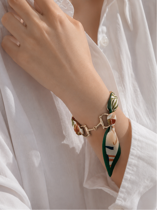 A close-up of a person's wrist wearing a gold-tone horsebit bracelet with two decorative silk ribbon charms in green, white, and red, styled with a white button-down shirt.