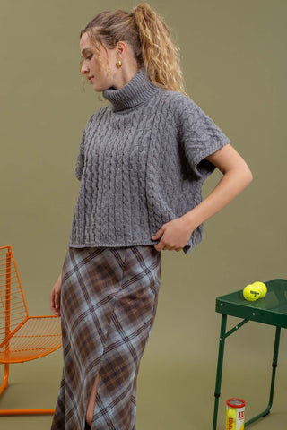 Woman wearing a gray cable knit sweater and plaid skirt against a green background
